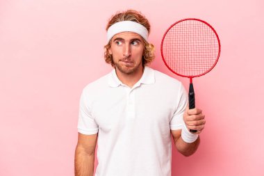 Pembe arka planda yalnız başına badminton oynayan beyaz bir adam kafası karışmış, kuşkulu ve güvensiz hissediyor..