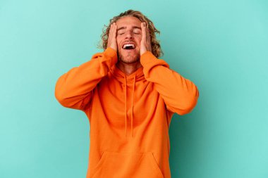 Mavi arka planda izole edilmiş genç beyaz adam ellerini başının üstünde tutarak neşe içinde gülüyor. Mutluluk kavramı.