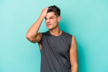 Kafkasyalı genç adam mavi arka planda izole edilmiş bir şekilde bir şeyi unutuyor. Alnına avuç içi ve gözlerini kapayarak vuruyor..