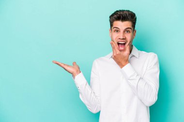 Mavi arka planda izole edilmiş genç beyaz bir adam avucunun içinde fotokopi alanı tutuyor, el üstünde tutuyor. Şaşırmış ve mutlu..