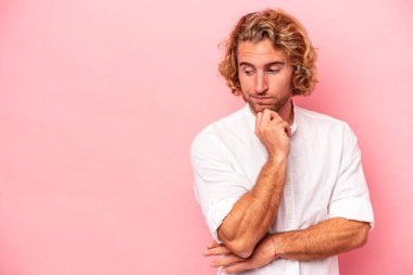Pembe arka planda izole edilmiş genç beyaz adam şüpheli ve şüpheci bir ifadeyle yana bakıyor..