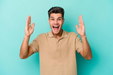 Mavi arka planda izole edilmiş genç beyaz adam hoş bir sürpriz alıyor, heyecanlı ve el kaldırıyor..
