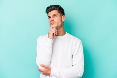 Kafkasyalı genç adam mavi arka planda izole edilmiş düşünüyor ve bakıyor, yansıtıyor, düşünüyor, bir fantezi kuruyor..