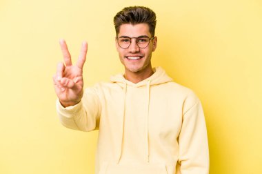 Sarı arka planda izole edilmiş genç beyaz adam zafer işareti gösteriyor ve gülümsüyor..