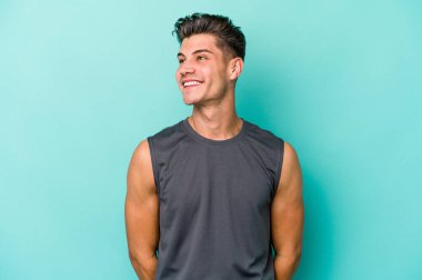 Kafkasyalı genç adam mavi arka planda izole edilmiş rahat ve mutlu bir şekilde gülüyor, boynunda dişlerini gösteriyor..