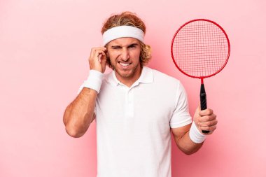 Badminton oynayan beyaz bir adam pembe arka planda izole edilmiş kulakları elleriyle kapatıyor..