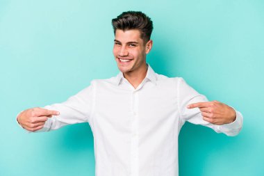 Mavi arka planda izole edilmiş genç beyaz adam bir gömlek fotokopi alanını işaret ediyor, gururlu ve kendinden emin.