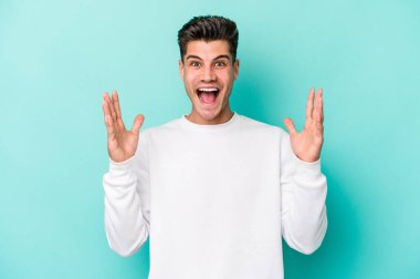 Mavi arka planda izole edilmiş genç beyaz bir adam zaferi ya da başarıyı kutluyor, şaşırıyor ve şok oluyor..