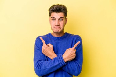 Sarı arka planda izole genç beyaz adam, iki seçenek arasında seçim yapmaya çalışıyor.