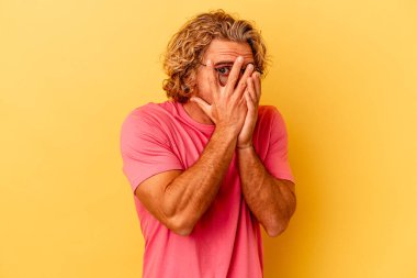 Sarı arka planda izole edilmiş genç beyaz adam parmaklarının arasından korkmuş ve gergin bir şekilde göz kırp..