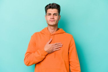 Kafkasyalı genç adam mavi arka planda izole edilmiş yemin ediyor, elini göğsüne koyuyor..
