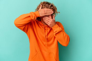 Mavi arka planda izole edilmiş genç beyaz adam kameraya parmaklarıyla göz kırpıyor, yüzünü kapatmaktan utanıyor..