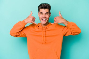 Mavi arka planda izole edilmiş genç beyaz adam parmaklarıyla bir cep telefonu hareketi gösteriyor..