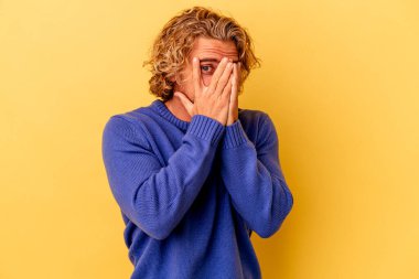 Sarı arka planda izole edilmiş genç beyaz adam parmaklarının arasından korkmuş ve gergin bir şekilde göz kırp..