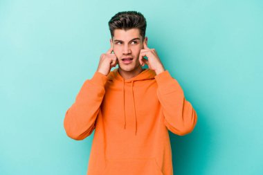 Genç beyaz adam mavi arka planda izole edilmiş parmaklarıyla kulaklarını kapatıyor, gürültülü bir ortam tarafından stresli ve umutsuz..