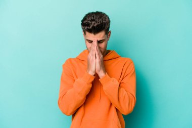 Kafkasyalı genç adam mavi arka planda izole edilmiş dua ediyor, bağlılık gösteriyor, dindar biri ilahi ilham arıyor..