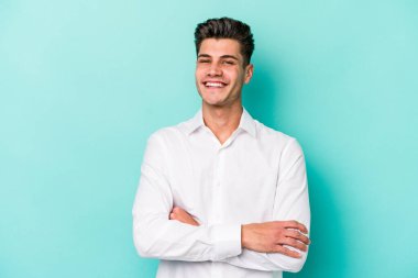 Mavi arka planda kendini güvende hisseden, kararlılıkla kollarını kavuşturan genç beyaz bir adam..