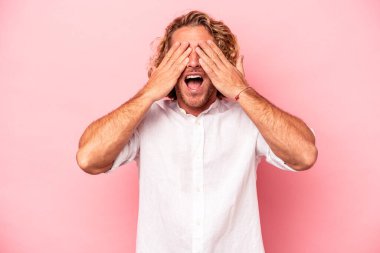 Pembe arka planda soyutlanmış genç beyaz adam elleriyle gözlerini kapatıyor, geniş bir gülümsemeyle bir sürpriz bekliyor..