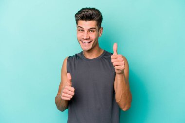 Kafkasyalı genç adam mavi arka planda izole edilmiş. Baş parmağını kaldırıyor, gülümsüyor ve kendinden emin..