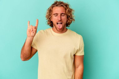 Mavi arka planda izole edilmiş genç beyaz adam parmaklarıyla kaya hareketi gösteriyor.