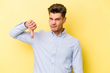 Sarı arka planda izole edilmiş genç beyaz adam baş parmağını aşağı gösteriyor, hayal kırıklığı konsepti..