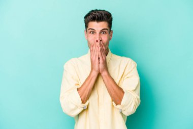 Mavi arka planda izole edilmiş genç beyaz adam elleriyle ağzını kapatırken şok oldu..