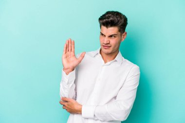 Kafkasyalı genç adam mavi arka planda izole, kafası karışmış, şüpheli ve güvensiz hissediyor..