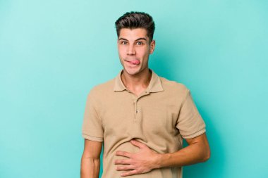 Mavi arka planda izole edilmiş genç beyaz adam karnına dokunuyor, nazikçe gülümsüyor, yemek yiyor ve tatmin oluyor..