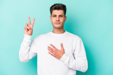 Kafkasyalı genç adam mavi arka planda izole edilmiş yemin ediyor, elini göğsüne koyuyor..