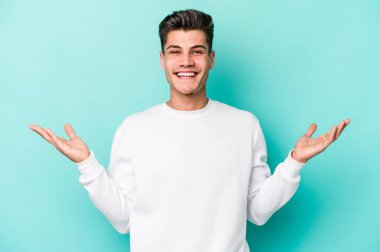 Mavi arka planda izole genç beyaz adam kollarıyla ölçek yapar, mutlu ve kendinden emin hisseder..