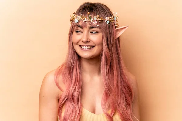 Young elf woman with pink hair isolated on beige background looks aside smiling, cheerful and pleasant.