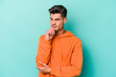 Kafkasyalı genç adam mavi arka planda izole edilmiş düşünüyor ve bakıyor, yansıtıyor, düşünüyor, bir fantezi kuruyor..