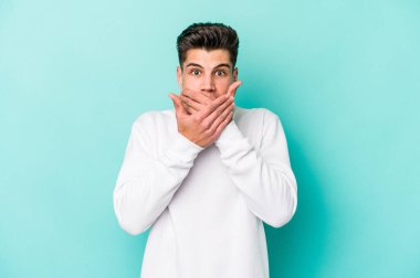 Mavi arka planda izole edilmiş genç beyaz adam elleriyle ağzını kapatırken şok oldu..