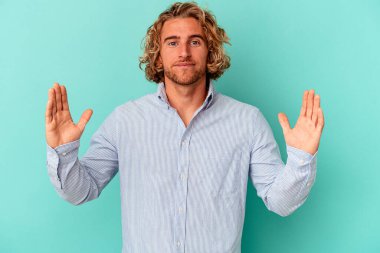 Kafkasyalı genç adam mavi arka planda izole edilmiş. Elinde işaret parmaklarıyla küçük bir şey tutuyor, gülümsüyor ve kendinden emin..