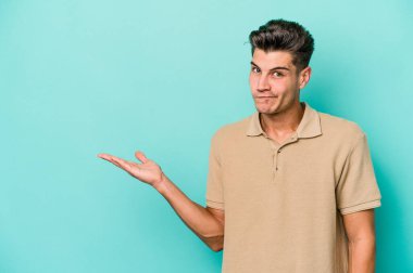 Mavi arka planda izole edilmiş genç beyaz bir adam bir palmiye üzerinde fotokopi alanı ve belinde bir el daha gösteriyor..