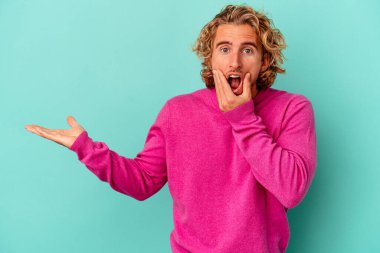 Mavi arka planda izole edilmiş genç beyaz bir adam avucunun içinde fotokopi alanı tutuyor, el üstünde tutuyor. Şaşırmış ve mutlu..