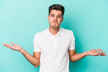 Kafkasyalı genç adam mavi arka planda izole edilmiş sorgulama jestinde şüphe ediyor ve omuz silkiyor..