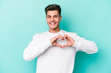Mavi arka planda izole edilmiş genç beyaz adam gülümsüyor ve elleriyle kalp şeklini gösteriyor..