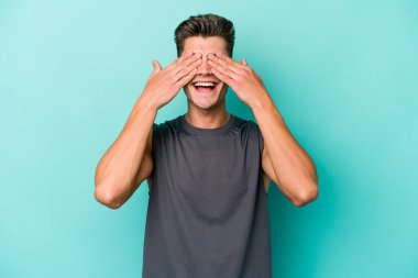 Mavi arka planda soyutlanmış genç beyaz bir adam elleriyle gözlerini kapatıyor, geniş bir gülümsemeyle bir sürpriz bekliyor..