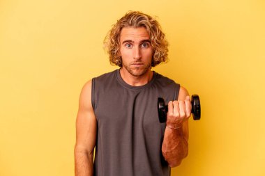 young sport man making weightlifting isolated on yellow background shrugs shoulders and open eyes confused. clipart