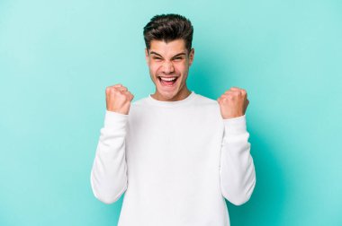 Kafkasyalı genç adam mavi arka planda izole edilmiş kaygısız ve heyecanlı tezahüratlar yapıyor. Zafer konsepti.