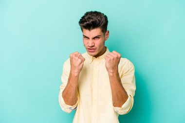 Mavi arka planda izole edilmiş genç beyaz adam yumruğunu kameraya gösteriyor, agresif yüz ifadesi var..