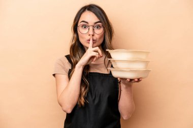 Beyaz tenli genç bir kadın bej arka planda saklanmış plastik kaplar taşıyor ya da bir sır saklıyor ya da sessizlik istiyor..