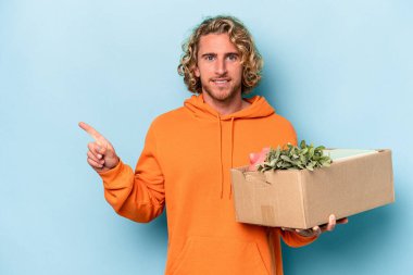 Kafkasyalı genç adam mavi arka planda izole edilmiş şeylerle dolu bir kutuyu kaldırırken hareket ediyor gülümsüyor ve boş alanda bir şeyler gösteriyor..