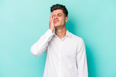 Kafkasyalı genç adam mavi arka planda izole edilmiş. Yorgun ve uykulu. Elini başının üstünde tutuyor..