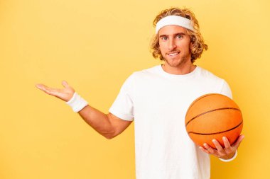 Young basketball player caucasian man isolated on yellow background showing a copy space on a palm and holding another hand on waist.