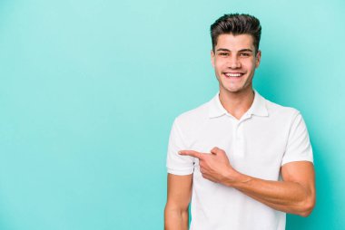 Mavi arka planda izole edilmiş genç beyaz adam gülümsüyor ve boş alanda bir şeyler gösteriyor..