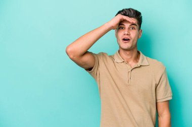 Kafkasyalı genç adam mavi arka planda izole edilmiş, uzaklara bakıyor ve elini alnına koyuyor..