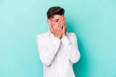 Kafkasyalı genç adam mavi arka planda izole edilmiş. Korkmuş ve gergin parmaklarıyla göz kırp..