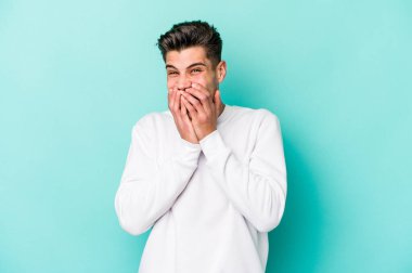 Mavi arka planda izole edilmiş genç beyaz bir adam bir şeye gülüyor, ağızlarını ellerle kapatıyor..